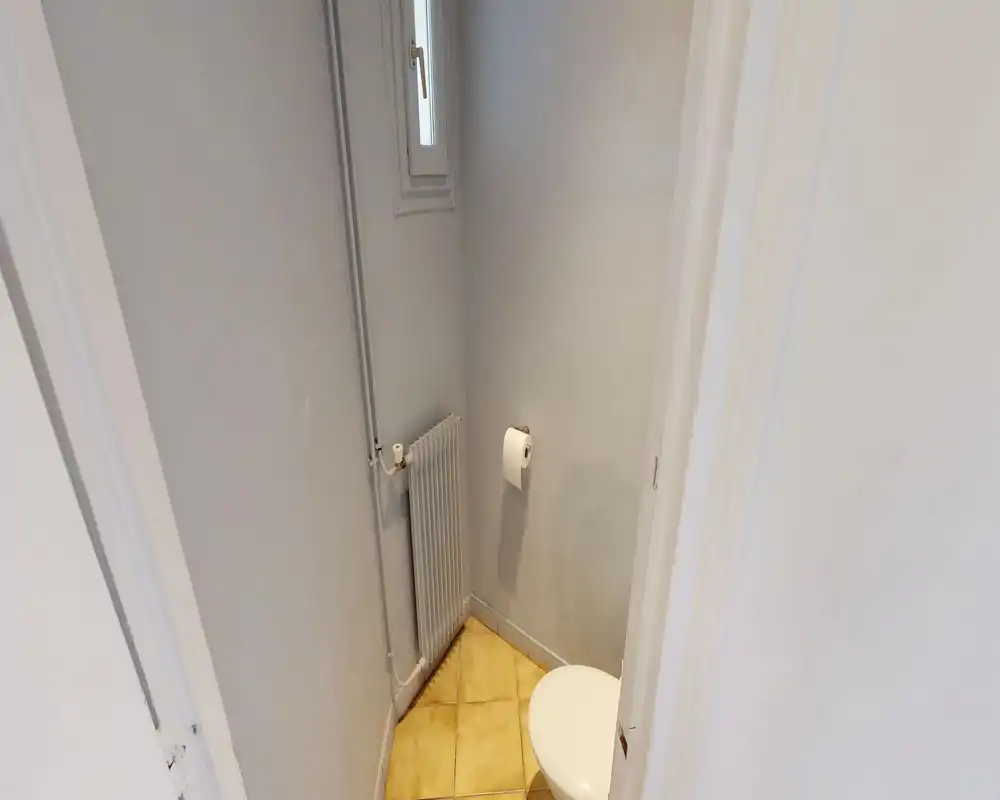 Small toilet room with a visible toilet seat, wall-mounted toilet paper holder and a vertical radiator; neutral light and simple finishes.