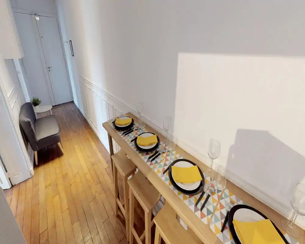 A narrow living/dining corridor with a long high table set for four, wooden stools and a small upholstered chair by the door; clean white walls and warm hardwood floors give a simple, modern look.