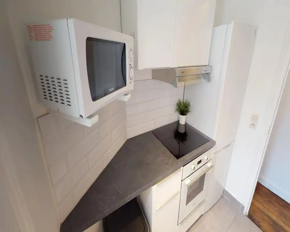 Compact modern kitchen with a ceramic cooktop, built-in oven, fridge and microwave. Clean white cabinetry and a dark stone-effect countertop create a tidy, functional space.