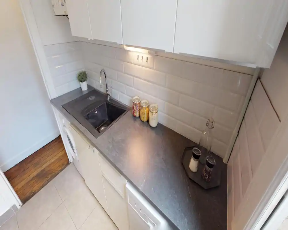 Compact modern kitchen with a dark countertop, integrated sink and white subway-tile backsplash; small decorative jars and under-cabinet lighting create a neat, functional workspace.