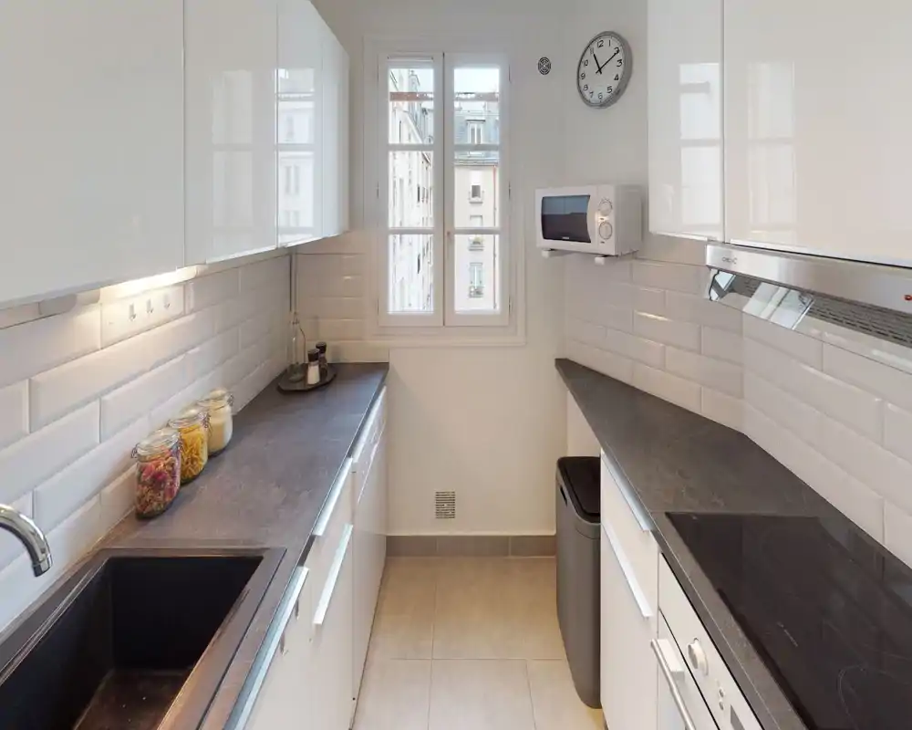 Bright, modern galley kitchen with dark countertops, white cabinetry, built-in stove and a window providing natural light. Clean, compact layout ideal for efficient cooking.