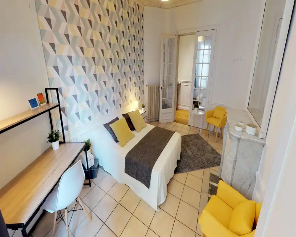 Bright, stylish bedroom with a double bed centered against a geometric accent wall, cozy textiles, a work desk, and two yellow armchairs that add cheerful accents.