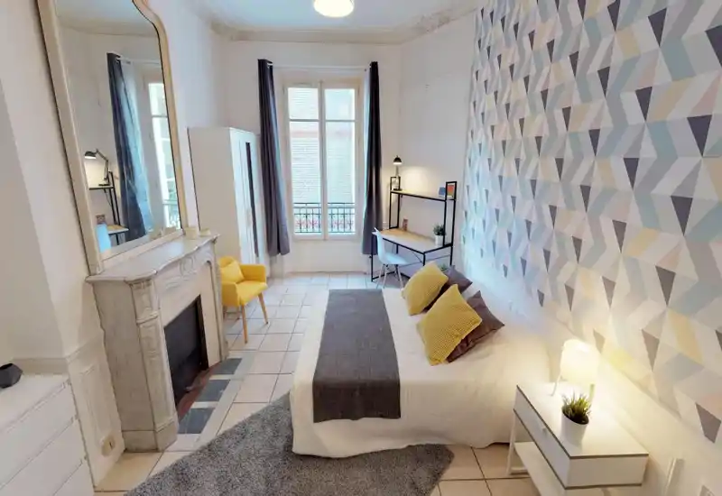 Bright, stylish bedroom with a comfortable double bed, decorative cushions, a bedside table with lamp, a work desk by the window and a patterned accent wall creating a modern, welcoming atmosphere.