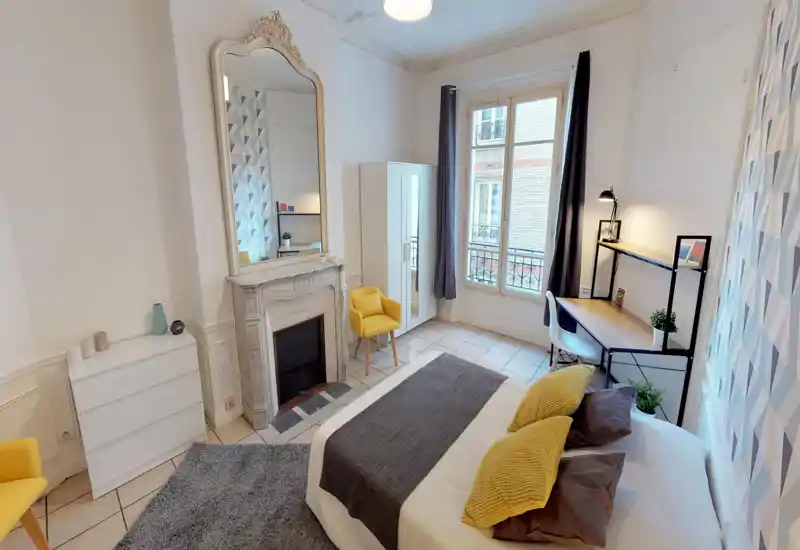 Bright, stylish bedroom with a double bed dressed in neutral linens and yellow accent pillows, a decorative fireplace with a large mirror, desk by the window, wardrobe and cozy rugs — ideal for a comfortable stay.