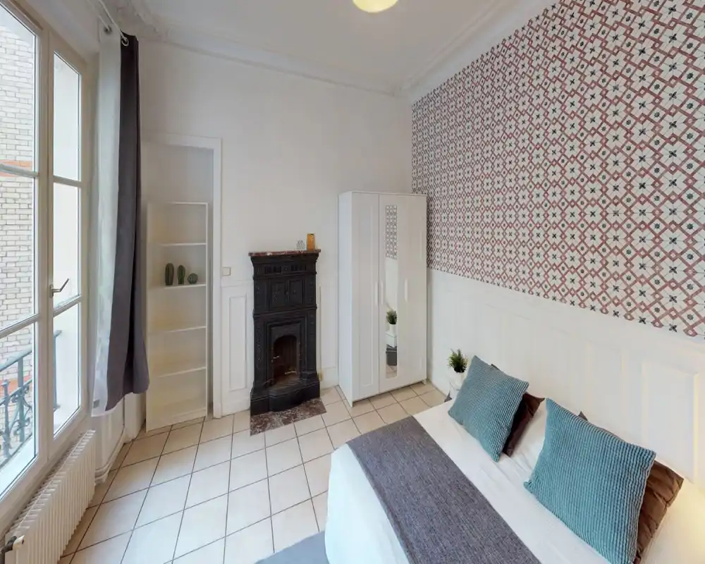 Bright, stylish bedroom with a double bed, decorative patterned wallpaper, white wardrobe and a classic fireplace; large windows provide plenty of natural light.