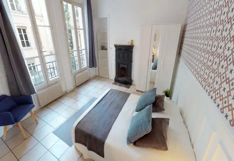 Spacious, bright bedroom with a large double bed, decorative pillows and a patterned accent wall. Tall windows provide abundant natural light and a small seating area adds comfort.