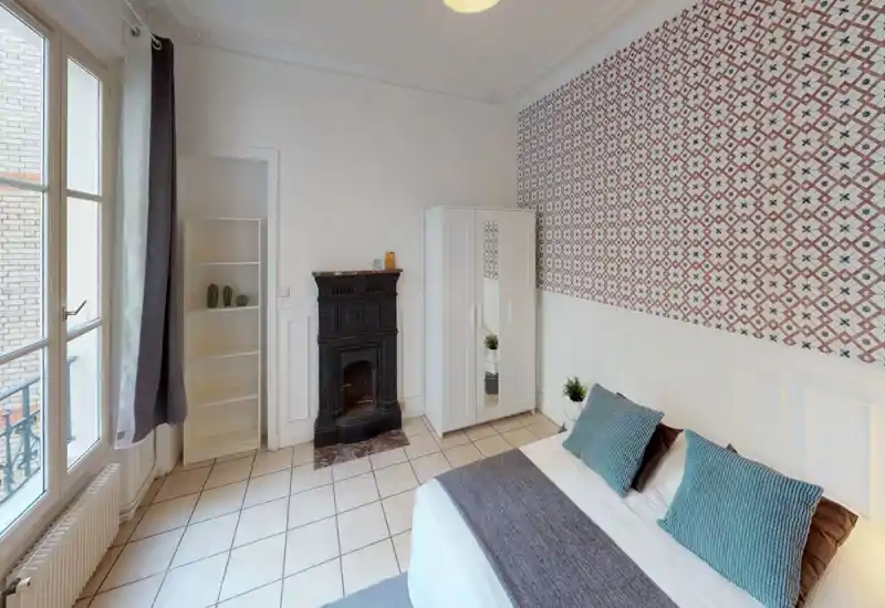 Bright, stylish bedroom with a double bed, decorative patterned wallpaper, white wardrobe and a classic fireplace; large windows provide plenty of natural light.