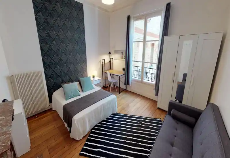 Bright, stylish bedroom with a double bed, patterned accent wall, wooden floors and a workspace by the window. The room includes a wardrobe and a sofa, offering comfortable living and practical storage.