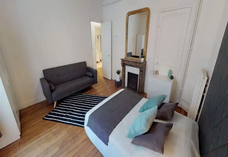 Well-styled bedroom with a comfortable double bed, decorative pillows and a soft throw; a small sofa and patterned rug add a cozy sitting area. Wood floors and a large gilded mirror above the fireplace give the room character and warmth.