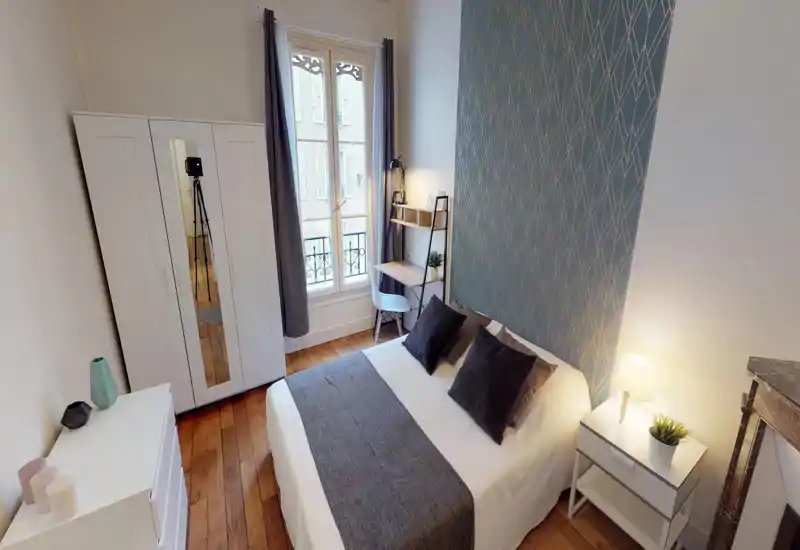 Bright, well-styled bedroom with a double bed, decorative cushions, bedside table with lamp, a wardrobe and a small desk by the window — ideal for city stays.