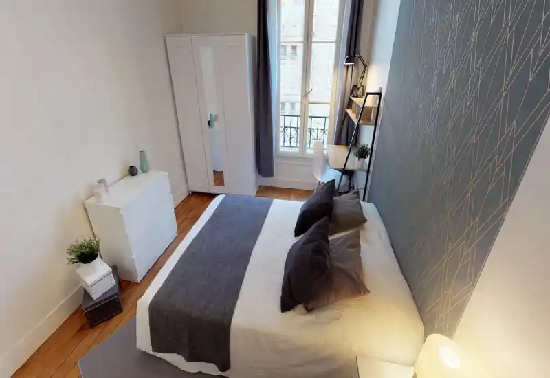 Bright, modern bedroom with a large double bed, decorative pillows and a textured feature wall. The room includes a wardrobe, dresser and small desk by the window, ideal for a comfortable stay.