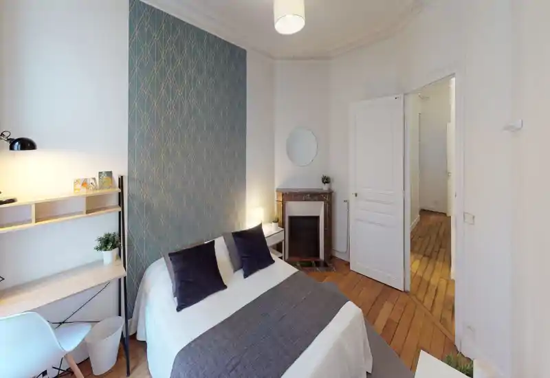 Bright, well-styled bedroom with a double bed, decorative pillows and a patterned accent wall; a desk area and bedside table complete the cozy layout.