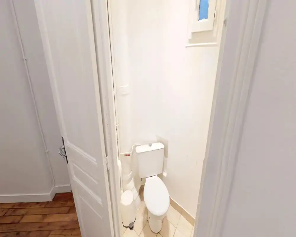Compact private WC with a close-coupled toilet, tiled floor and simple white walls; small waste bin and visible plumbing make it a functional guest bathroom.