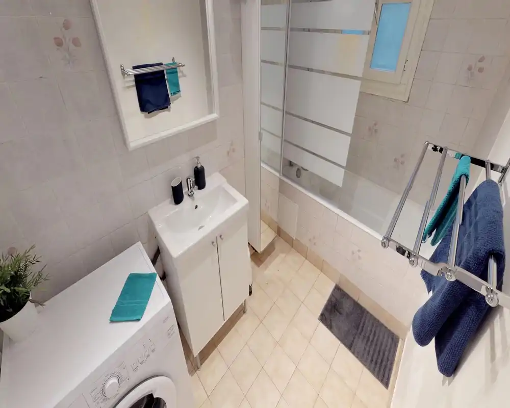 Clean, compact bathroom with a sink cabinet, bathtub with glass screen, washing machine and towel racks; neutral tiles and good lighting make it practical for guests.