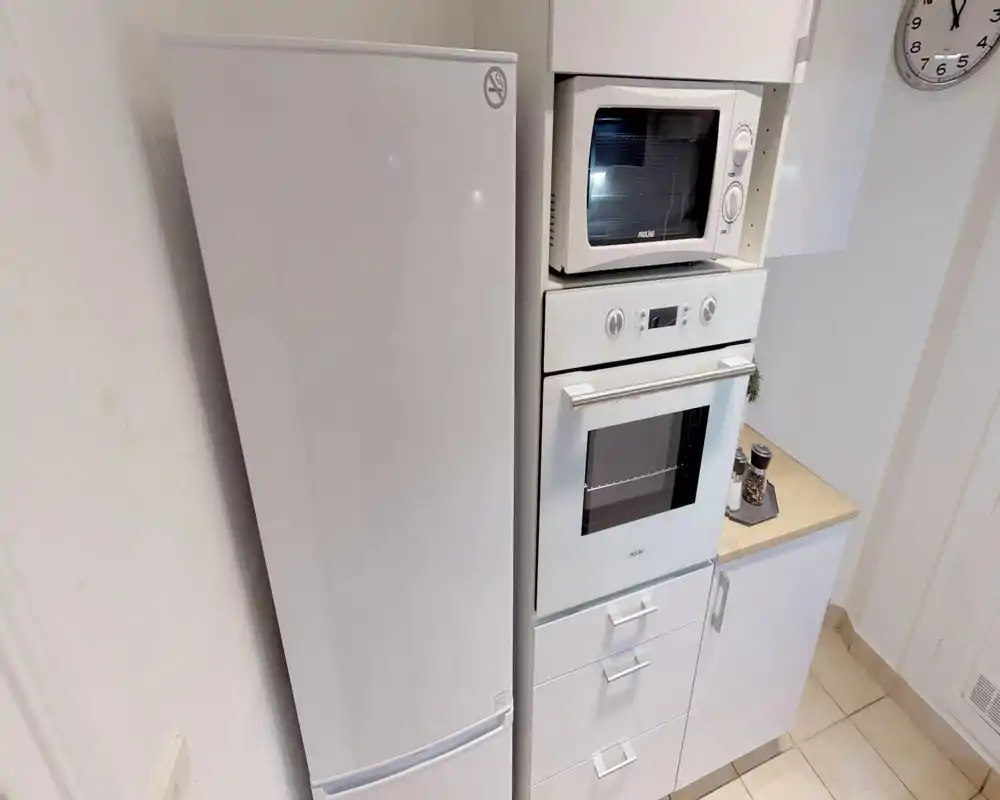 Compact modern kitchen corner with white fridge, built-in oven and microwave, and a tidy countertop with spices — bright and clean layout ideal for short-term rentals.