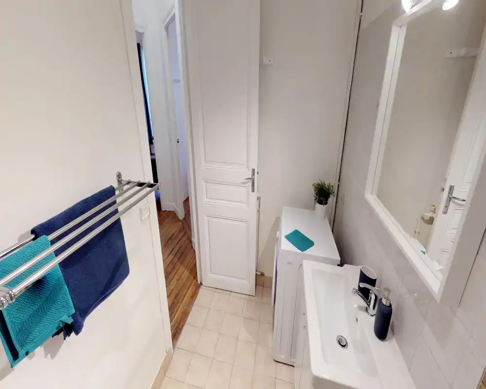 Clean, well-lit bathroom with a white sink, mirror, towel rack and tiled floor; small washing machine or cabinet next to the sink and decorative plant add a fresh touch.