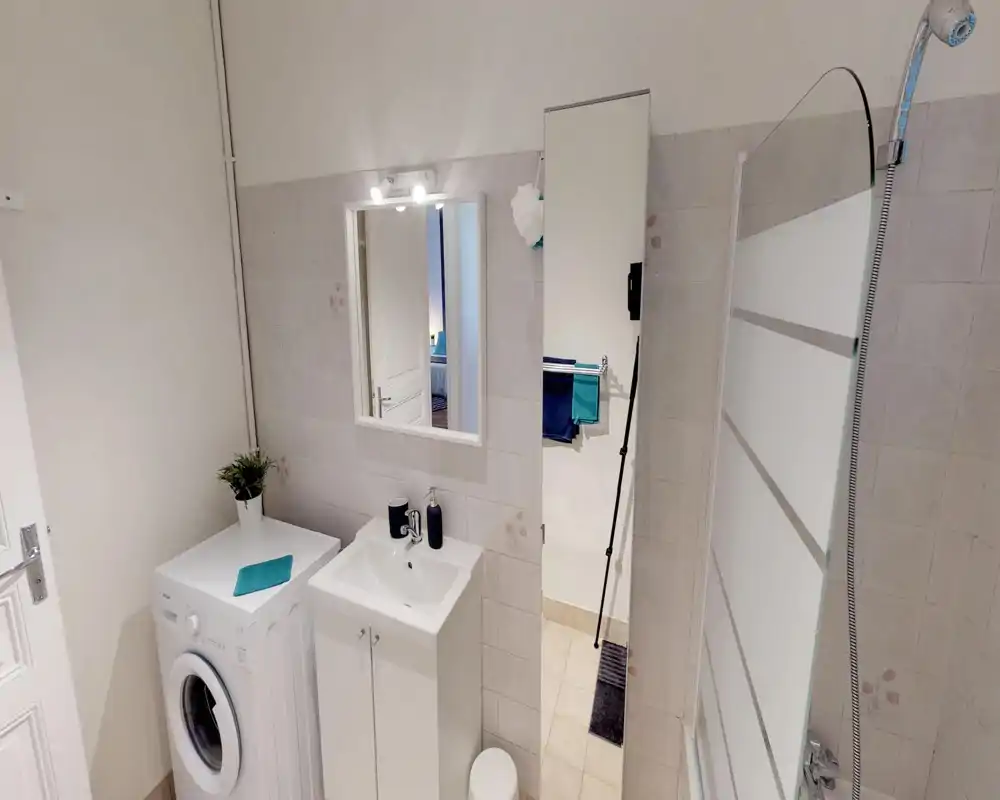 Clean, compact bathroom with a sink cabinet, mirror, shower screen and a washing machine. Neutral tiles and bright lighting create a tidy, functional space.