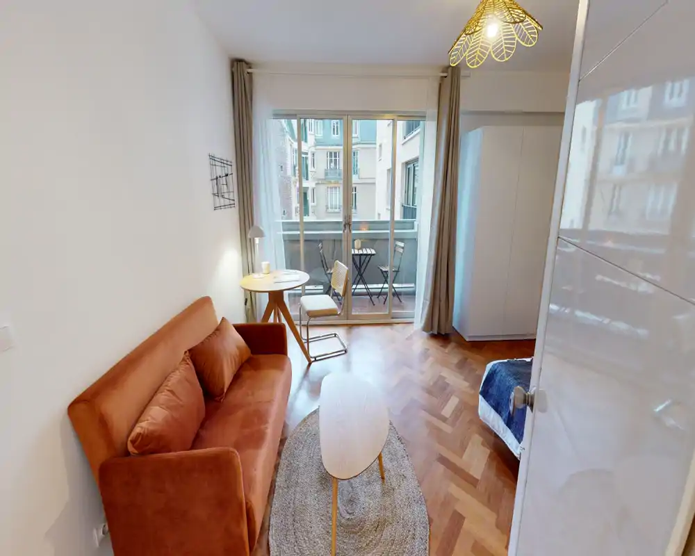 Chambre-studio lumineuse et chaleureuse avec un canapé couleur rouille, petite table près du balcon et lit partiellement visible — parfaite pour une personne. Grandes portes vitrées donnant sur un balcon apportent beaucoup de lumière naturelle.