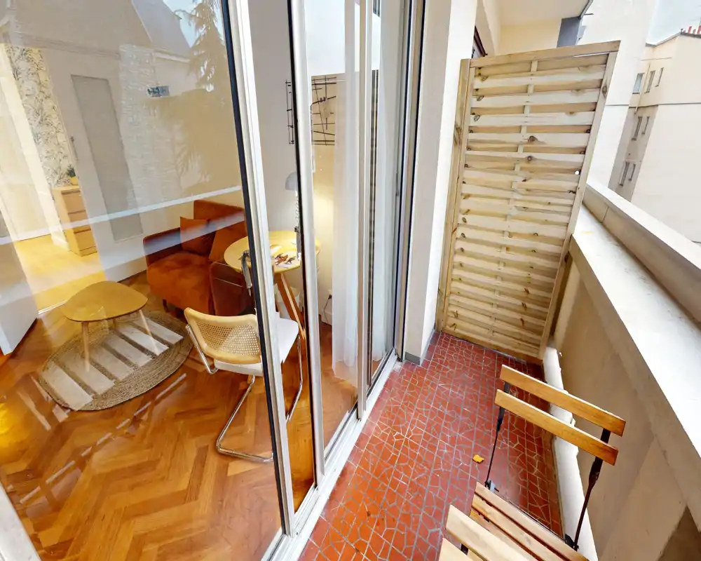 Petit balcon urbain avec sol carrelé rouge, chaises pliantes en bois et écran d'intimité ; vue sur un salon cosy à travers de grandes portes-fenêtres coulissantes.