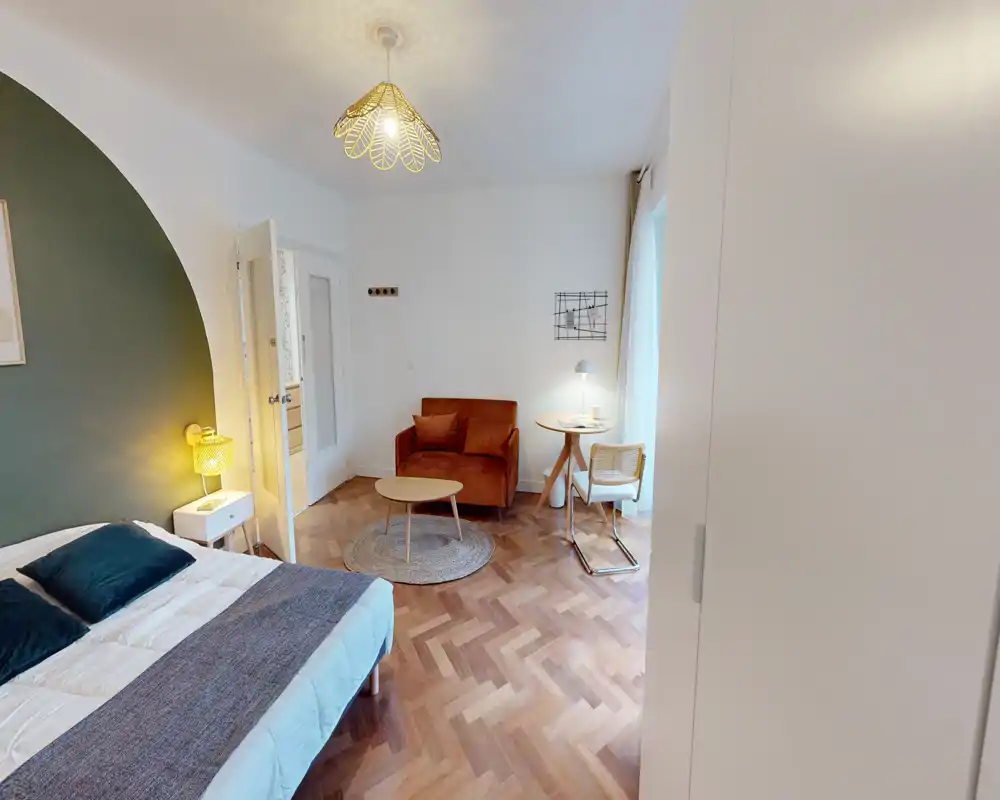 Chambre lumineuse et élégante avec lit double, textiles chaleureux et un coin salon cosy composé d’un fauteuil et d’une petite table — idéale pour se détendre ou travailler près de la fenêtre.