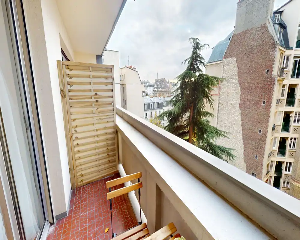 Balcon étroit et ensoleillé avec sol carrelé, petite chaise pliante et vue sur la ville — parfait pour un café le matin ou une pause en plein air.