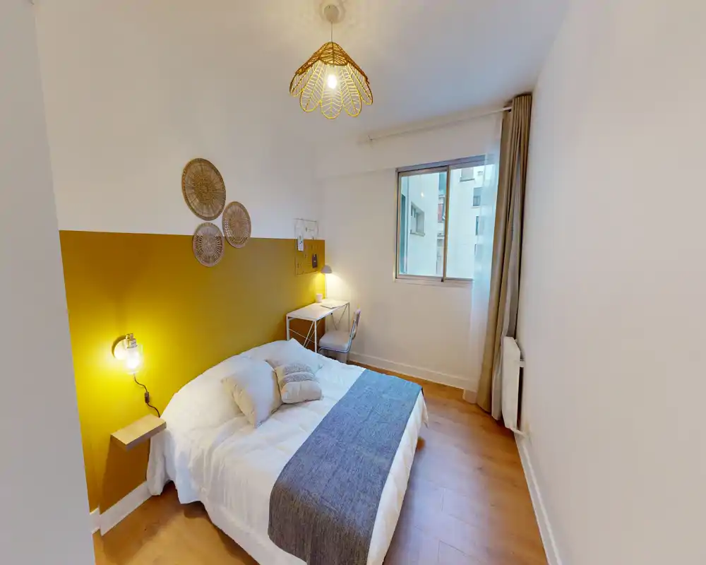 Chambre lumineuse et décorée avec lit complet, mur d'accent moutarde et lampes de chevet. La pièce comprend un petit bureau près de la fenêtre et un sol en bois pour une ambiance chaleureuse et moderne.