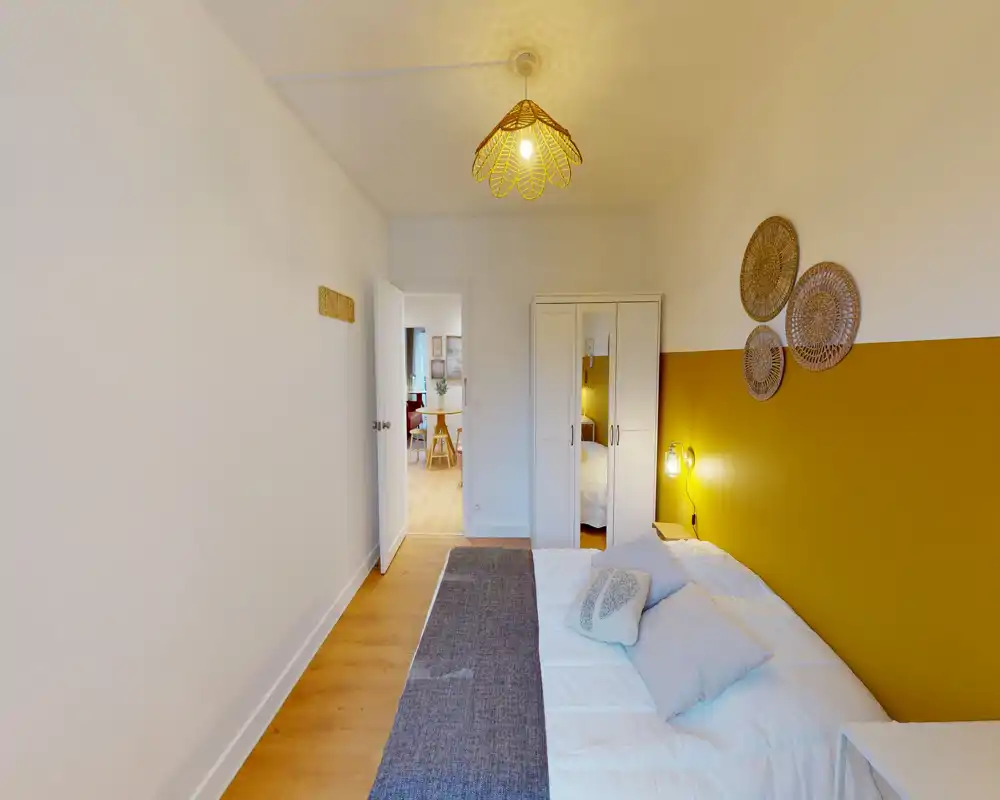 Chambre lumineuse et cosy avec un lit double confortable contre un mur d'accent jaune moutarde, ornements muraux tressés et une armoire miroir. Cliché net avec une lumière chaude, idéal pour une annonce locative.
