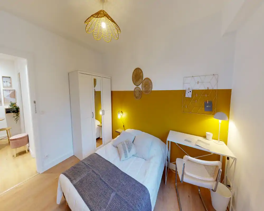 Chambre lumineuse et bien décorée avec lit double, lampes de chevet, armoire miroir et petit bureau — mur d'accent ocre et sol en bois pour une ambiance moderne et chaleureuse.