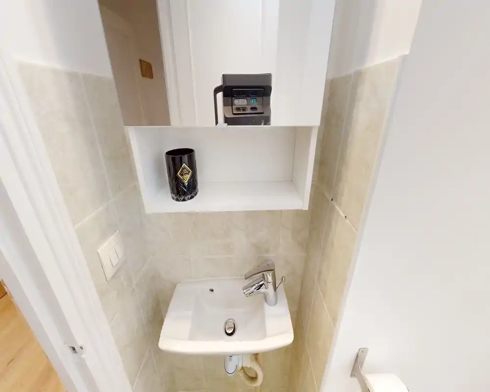 Coin lavabo compact avec armoire murale, petit lavabo en céramique et robinetterie chrome moderne; carrelage clair et petite étagère pour produits d'hygiène.