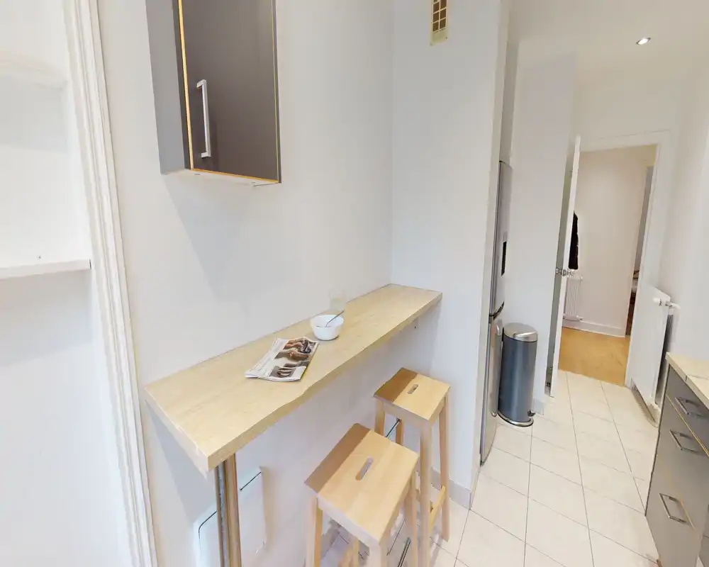 Cuisine compacte et moderne avec un bar en bois et deux tabourets, meubles neutres et sol carrelé — lumineuse et pratique pour repas rapides.