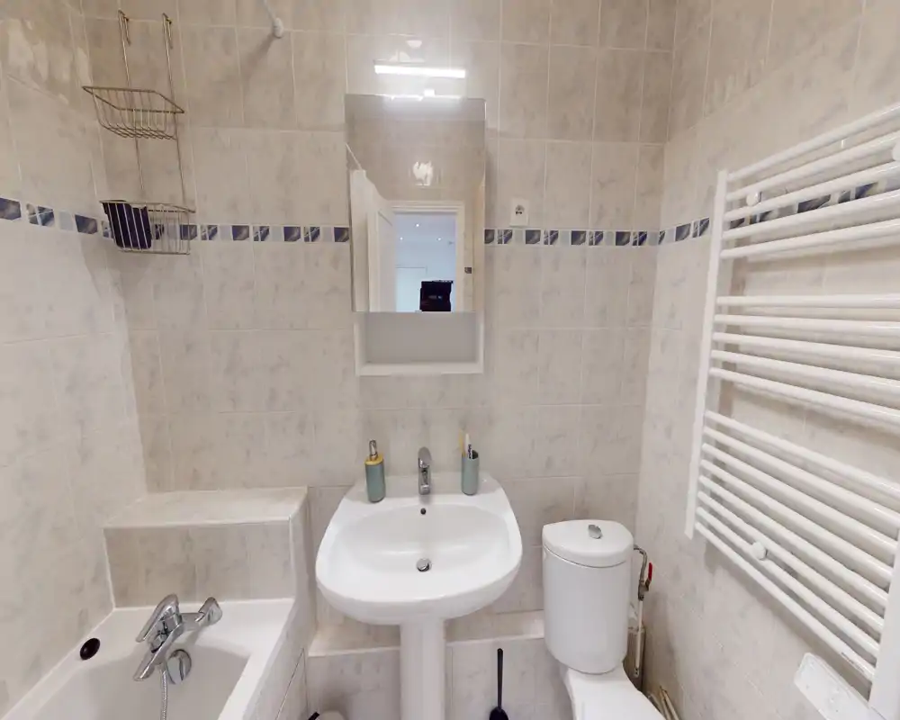 Salle de bain lumineuse et compacte avec baignoire, lavabo sur colonne, WC et radiateur sèche-serviettes mural. Carrelage propre, petit miroir-meuble et rangement discret pour un usage pratique.