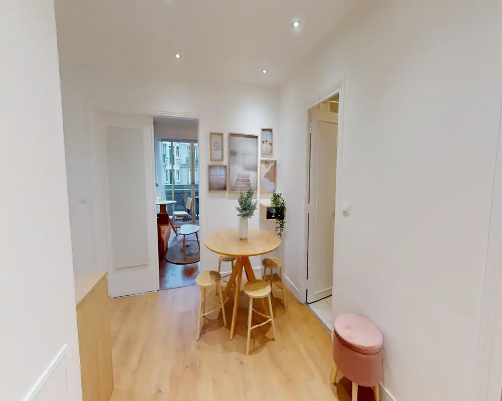 Coin repas compact avec table ronde en bois, trois tabourets et décorations douces ; sol chaleureux et cadres muraux créent un espace repas cosy.