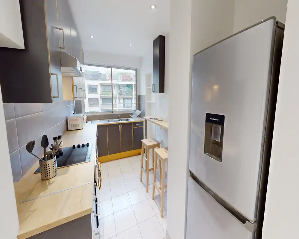 Cuisine moderne en enfilade avec plans de travail en bois, plaque de cuisson intégrée et réfrigérateur en inox. Fenêtre lumineuse au fond et deux tabourets en bois pour un espace de cuisson compact et fonctionnel.