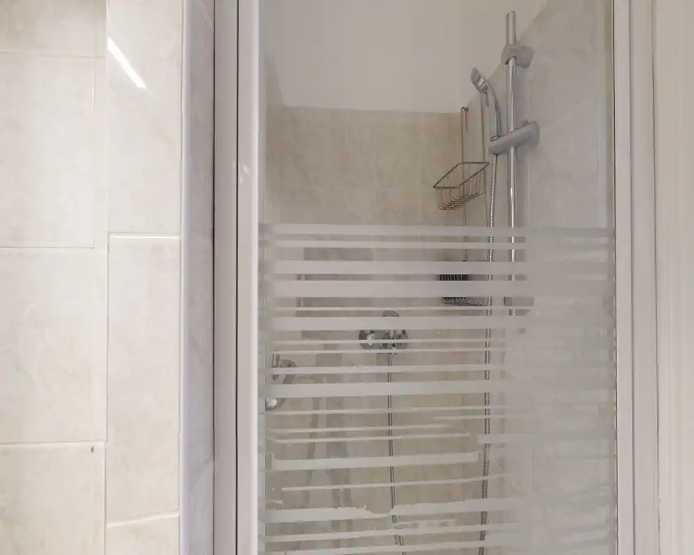 Douche carrelée et lumineuse avec porte en verre et pommeau de douche chromé visible ; détail de salle de bain propre et minimal, idéal pour les annonces locatives.