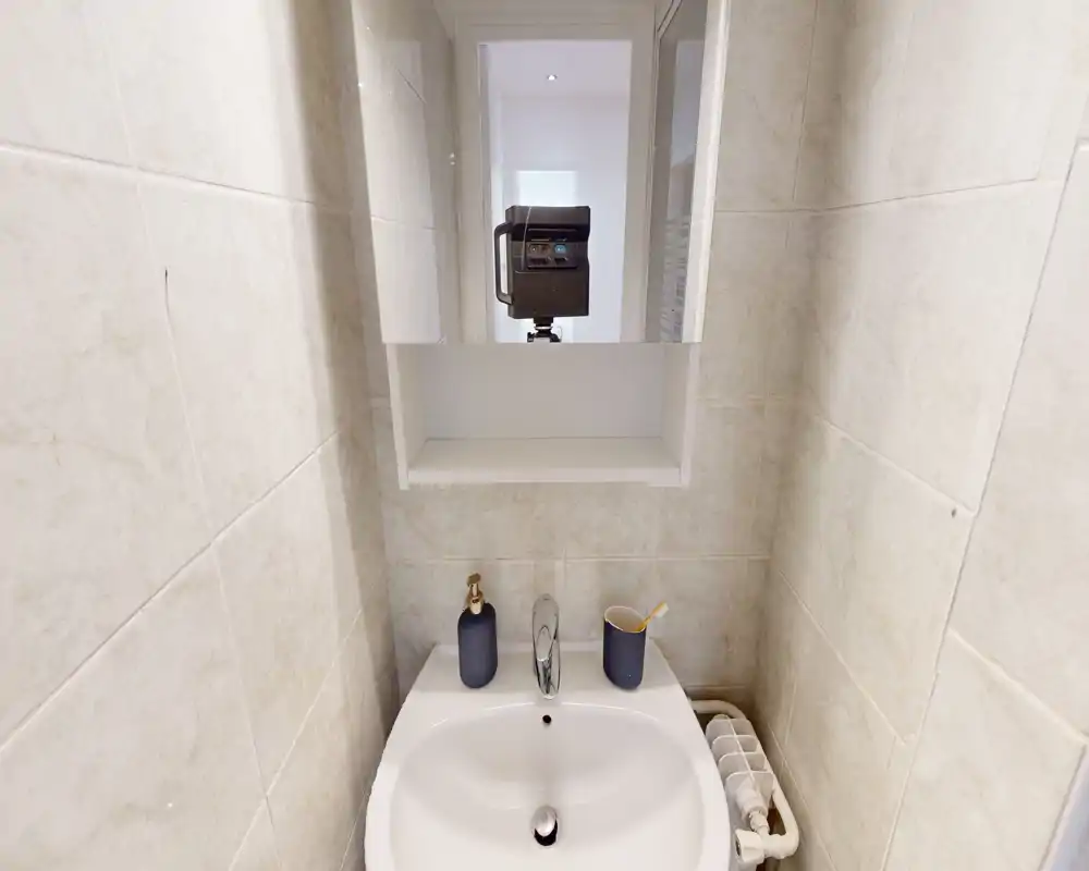 Petit espace lavabo de salle de bain bien éclairé avec vasque en céramique blanche, robinet chromé, miroir armoire et quelques accessoires assortis pour une impression soignée.