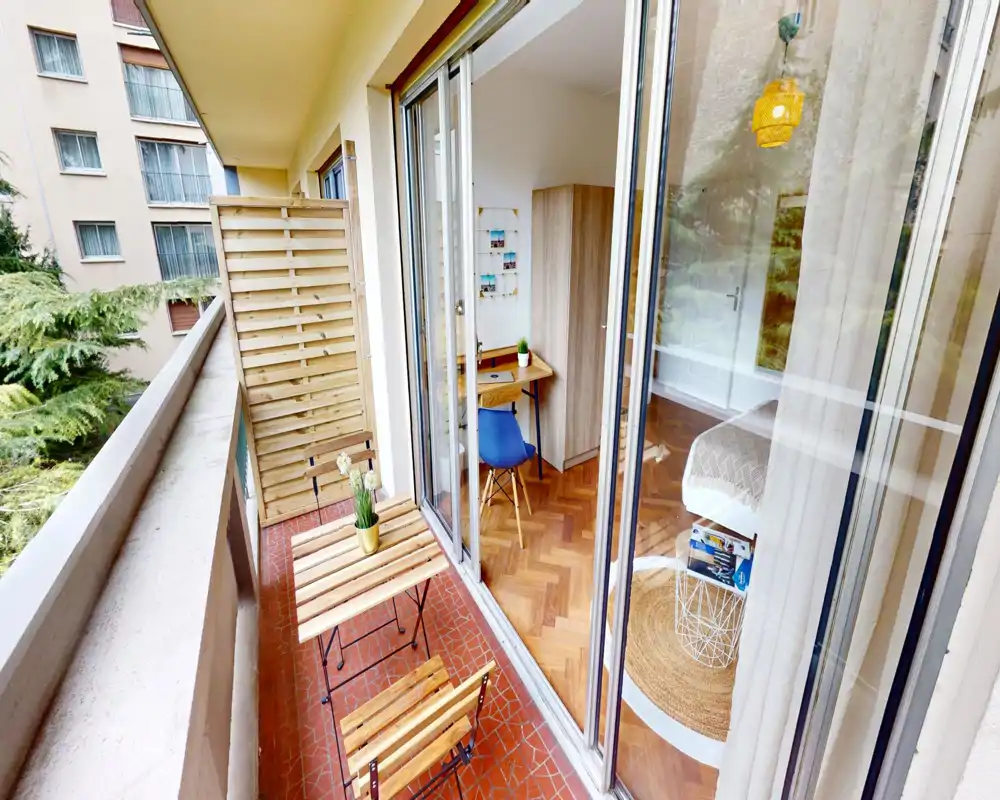 Chambre moderne et lumineuse aperçue derrière de grandes portes-fenêtres coulissantes, avec tapis cosy, table de chevet et un bureau; un petit balcon avec mobilier en bois est au premier plan.