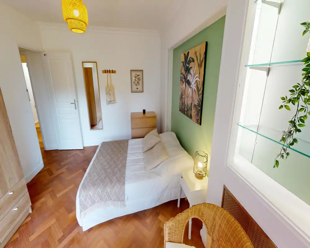 Chambre lumineuse et soignée avec un lit double, linge doux, table de chevet avec lampe, fauteuil en osier et une œuvre murale sur un mur vert.