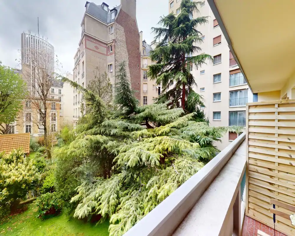 Balcon ensoleillé donnant sur une cour verdoyante avec de grands arbres et des immeubles voisins — idéal pour prendre un café le matin ou se détendre dehors.