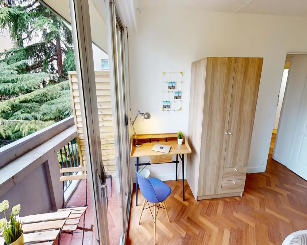 Espace de vie lumineux et compact avec un petit bureau près des portes-fenêtres s'ouvrant sur le balcon ; parquet et armoire forment un coin chaleureux et fonctionnel, parfait pour le télétravail.