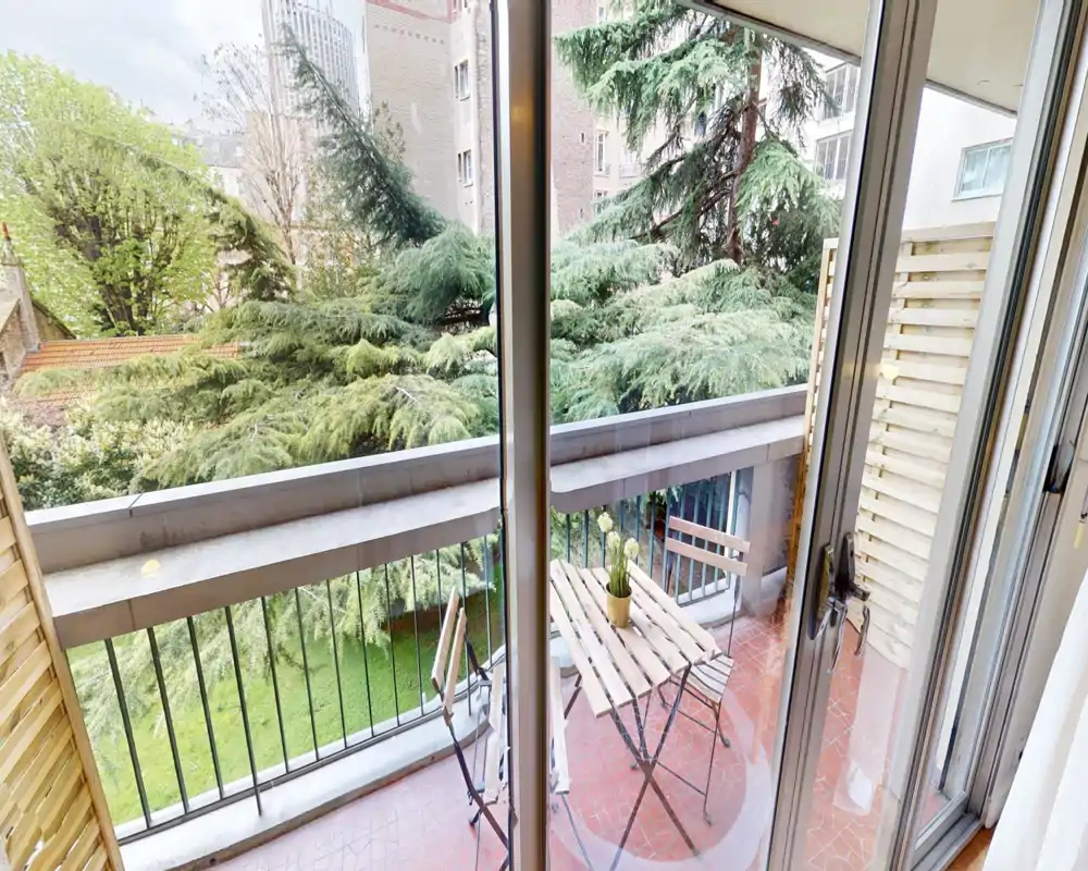 Small furnished balcony with a bistro table and two chairs opening onto a green courtyard—ideal for morning coffee with pleasant tree views.