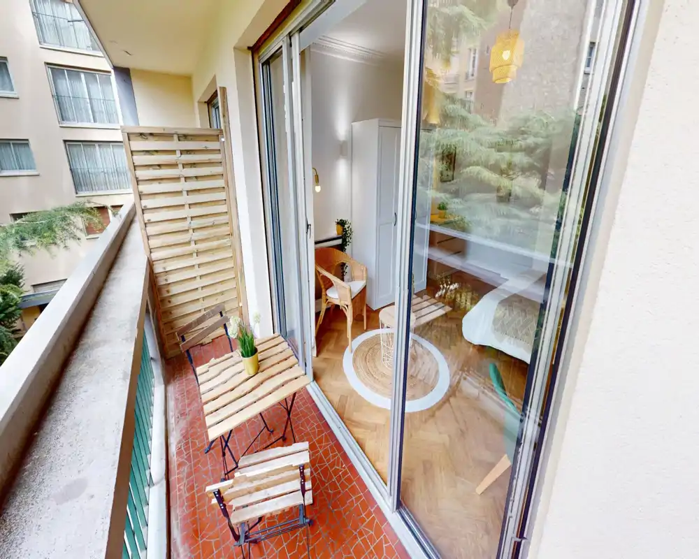 Bright bedroom viewed from a balcony with sliding glass doors; tasteful wooden flooring, a cozy chair, round rug and a neatly made bed visible through the glass.
