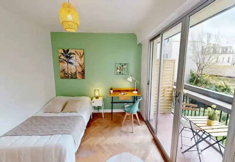Sunny, well-staged bedroom with a comfortable double bed, bedside table and a small desk next to large sliding doors opening onto a balcony. The green accent wall and natural light create a bright, inviting space ideal for relaxing or studying.