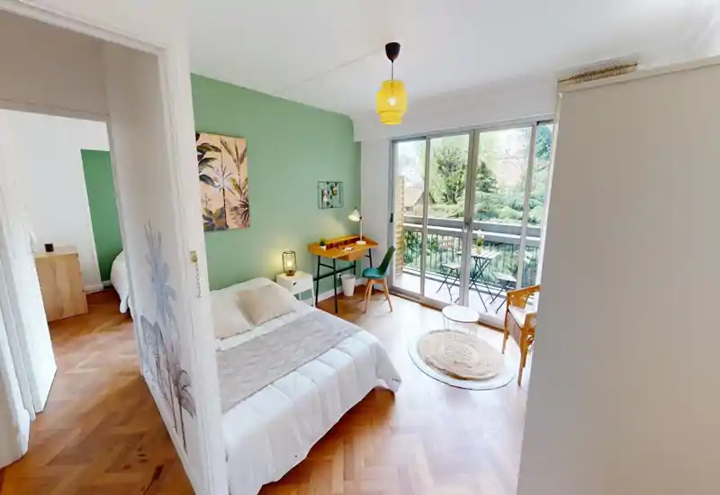 Bright, stylish bedroom with a king/queen bed, bedside table and a small desk by sliding glass doors opening to a balcony with outdoor seating — warm green accent wall and wooden parquet floors add character.