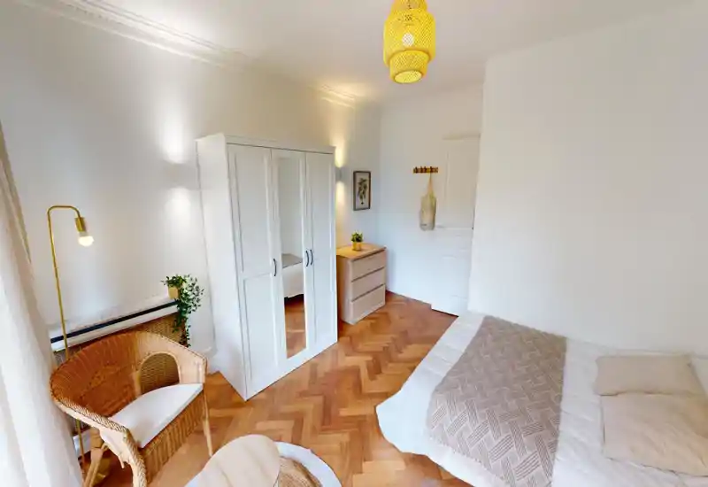 Bright, cozy bedroom with a comfortable double bed, white wardrobe with mirrored door, wooden parquet flooring and rattan seating — ideal for restful stays.