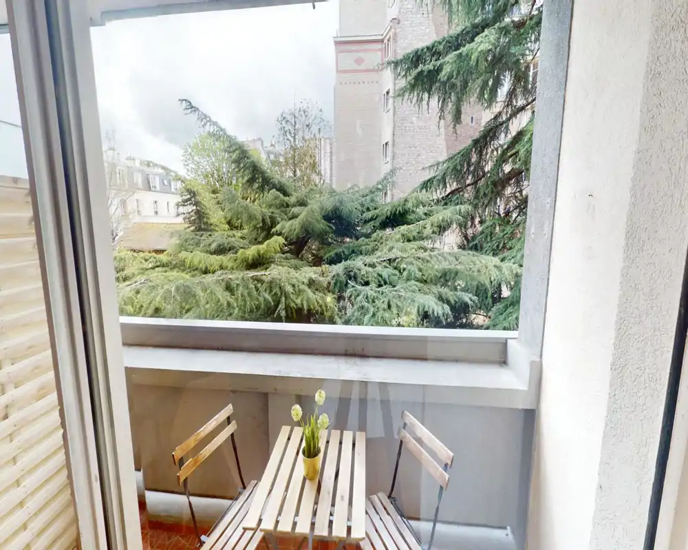 Piccolo balcone soleggiato con tavolino pieghevole in legno e due sedie, affacciato su alberi maturi e edifici vicini — perfetto per il caffè mattutino.