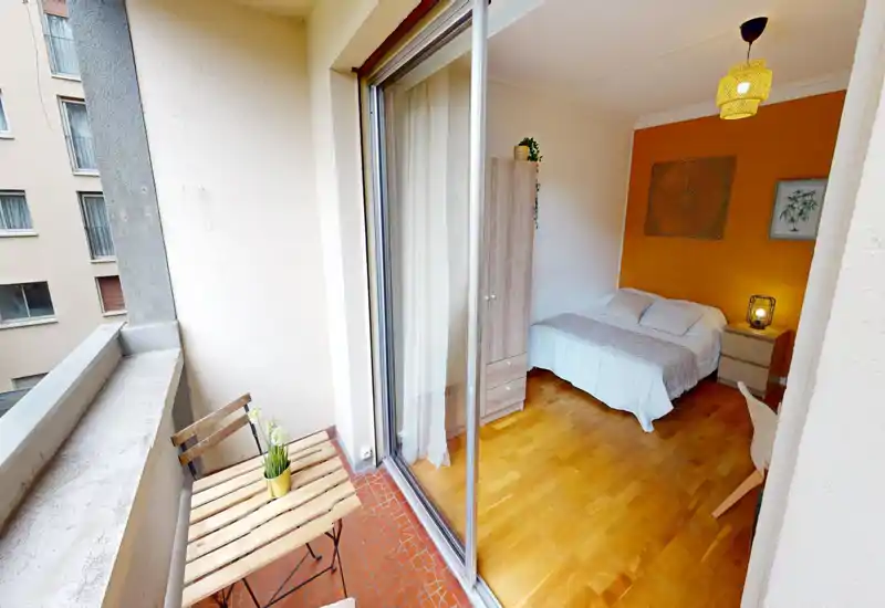 Camera da letto luminosa con parete color pesca, letto matrimoniale, comodino e armadio; ampia porta scorrevole in vetro che si apre su un piccolo balcone con tavolino.