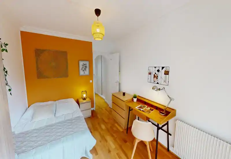 Camera da letto luminosa e accogliente con un grande letto comodo, parete accentuata color ocra, lampada da comodino e scrivania in legno ordinata — perfetta per riposare e lavorare.