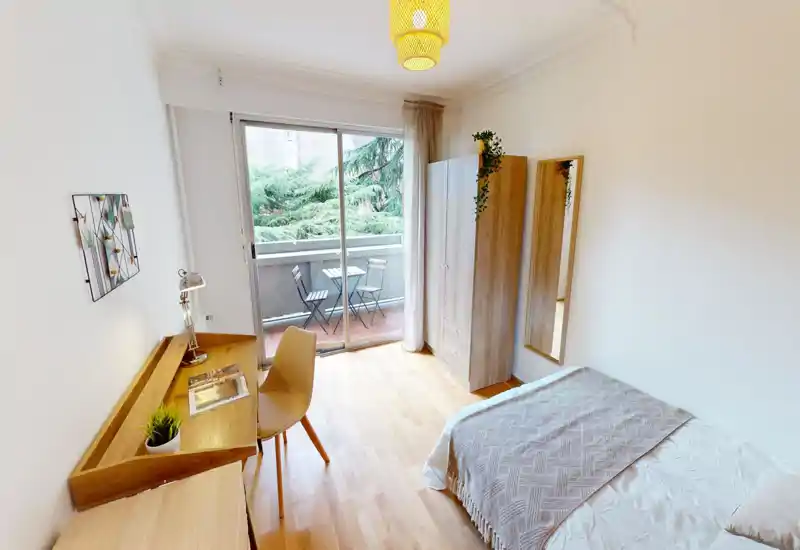 Camera da letto luminosa e moderna con letto ben rifatto, armadio e scrivania in legno, e porta scorrevole che si apre su un piccolo balcone con sedute esterne.