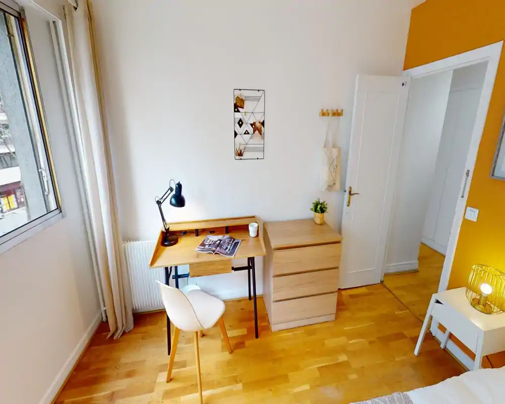 Bright, neatly styled bedroom corner with a wooden desk, chair and chest of drawers; warm accents and natural light create a welcoming study area beside the bed.