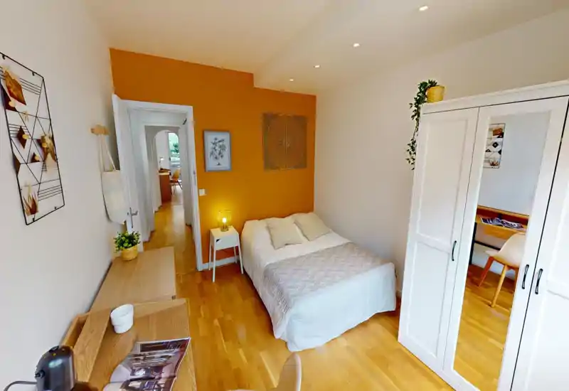 Bright, stylish bedroom with a double bed against a warm ochre accent wall, bedside table and lamp, desk area and a mirrored wardrobe — cozy and well lit for rental photos.
